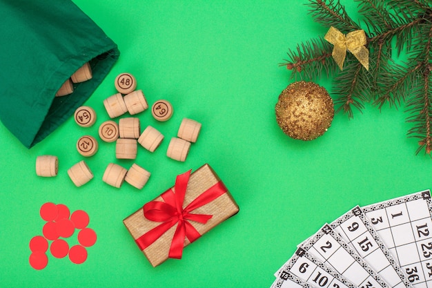 Juego de mesa de lotería. Barriles de madera de lotería con bolsa, tarjetas de juego y fichas rojas para un juego de lotería, rama de abeto de Navidad, pelota de juguete y caja de regalo sobre fondo verde. Vista superior