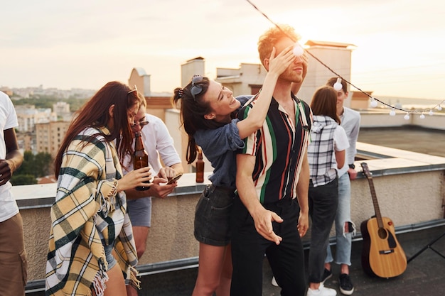 Juego Los ojos del hombre están cubiertos por las manos Un grupo de jóvenes con ropa informal tienen una fiesta en la azotea juntos durante el día