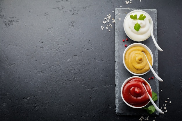 Juego de tres salsas: mayonesa, mostaza y ketchup en tazones de cerámica blanca sobre piedra negra u hormigón.