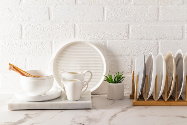 Un juego de utensilios de cocina sobre una encimera de mármol blanco, una secadora con placas contra el fondo de una pared de ladrillo blanco, una casa ecológicamente limpia