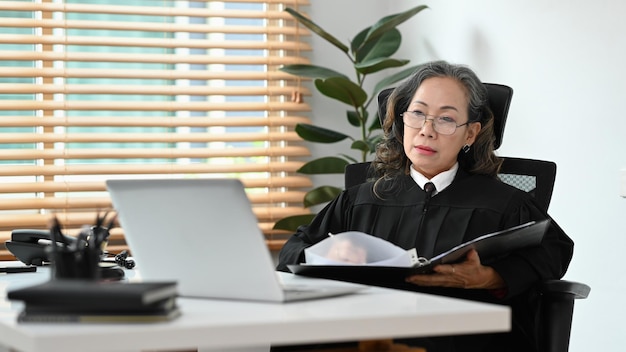 Juez o abogada madura que lee un documento de derecho judicial y usa una computadora portátil en su lugar de trabajo