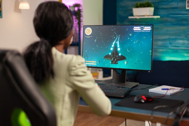 Jugador africano profesional que juega al tirador espacial con joystick inalámbrico por la noche. La mujer competitiva del jugador cibernético que realiza el torneo de videojuegos utiliza un joystick profesional.