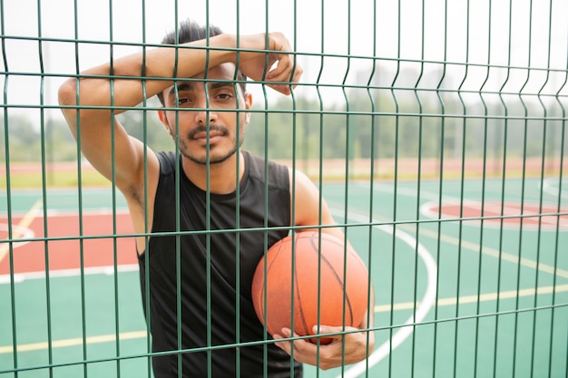 Jugador de baloncesto masculino serio con bola de pie contra la red mientras te mira en el descanso en el patio de recreo