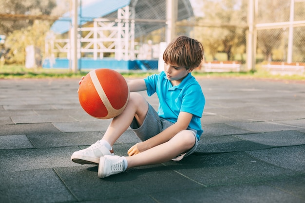 Foto un jugador de baloncesto de niño con una camiseta azul está sentado en la cancha de baloncesto con una pelota
