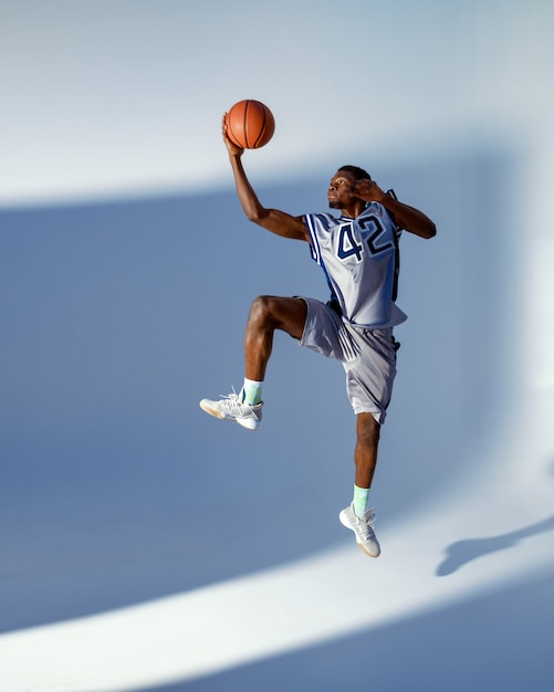 El jugador de baloncesto con pelota muestra su habilidad en el estudio, salto de altura en acción, fondo de neón. Baller masculino profesional en ropa deportiva jugando al deporte, deportista alto