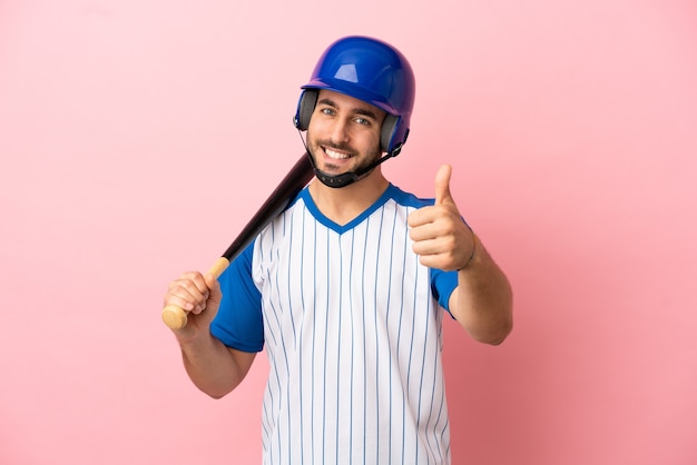 Jugador de béisbol con casco y bate aislado sobre fondo rosa con pulgares hacia arriba porque ha sucedido algo bueno