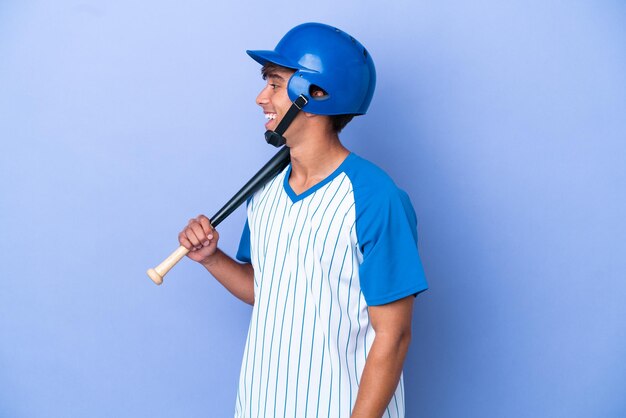 Foto jugador de béisbol caucásico con casco y bate aislado sobre fondo azul riendo en posición lateral