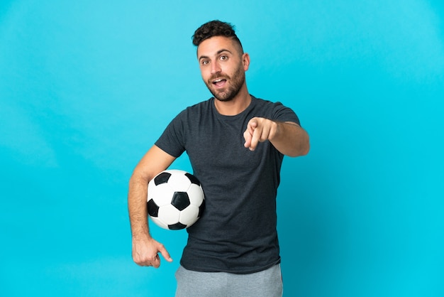 Jugador de fútbol aislado en la pared azul sorprendido y apuntando hacia el frente