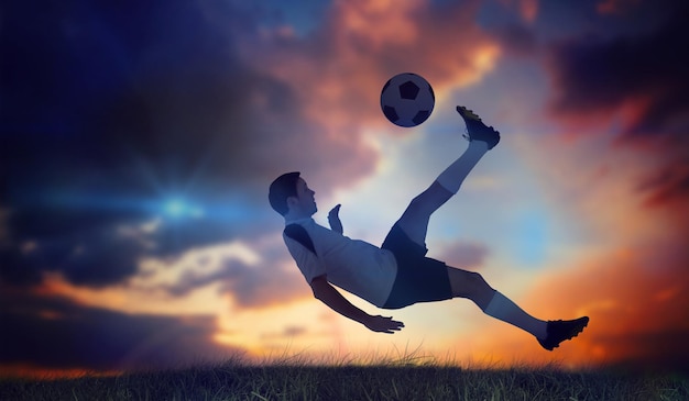 Jugador de fútbol de blanco pateando hierba verde bajo un cielo azul oscuro y naranja