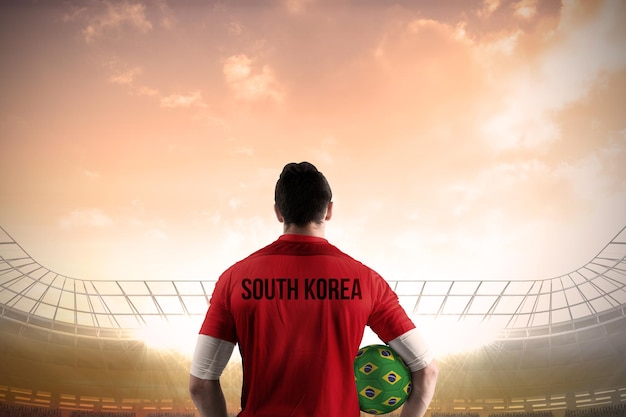 Jugador de fútbol de corea del sur sosteniendo la pelota contra un gran estadio de fútbol bajo un cielo azul nublado