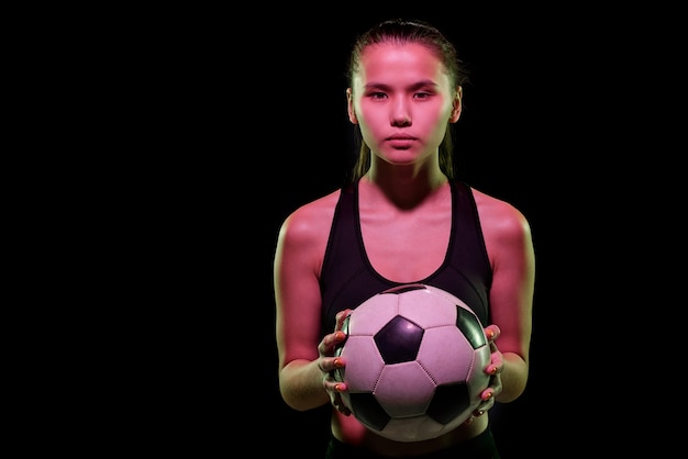 Jugador de fútbol femenino bastante joven que sostiene el balón de fútbol mientras entrena en aislamiento delante de la cámara sobre fondo negro