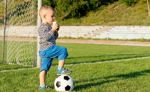 Jugador de fútbol de niño de pie con el pie en la pelota bebiendo jugo en la luz del atardecer mientras toma un descanso de jugar