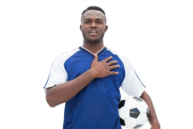 Jugador de fútbol en pie azul con la pelota escuchando el himno