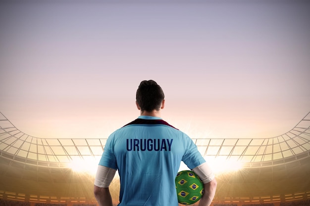 Jugador de fútbol de Uruguay sosteniendo la pelota contra un gran estadio de fútbol bajo un cielo azul