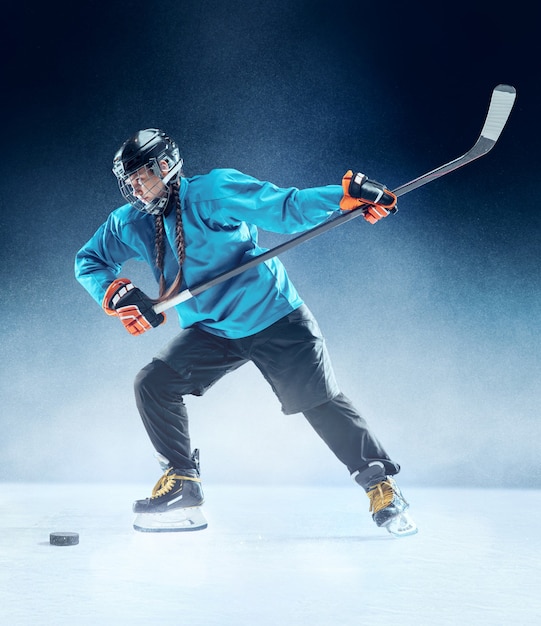 Jugador de hockey femenino joven con el palo en la cancha de hielo y fondo azul. Deportista con equipo y casco practicando. Concepto de deporte, estilo de vida saludable, movimiento, movimiento, acción.