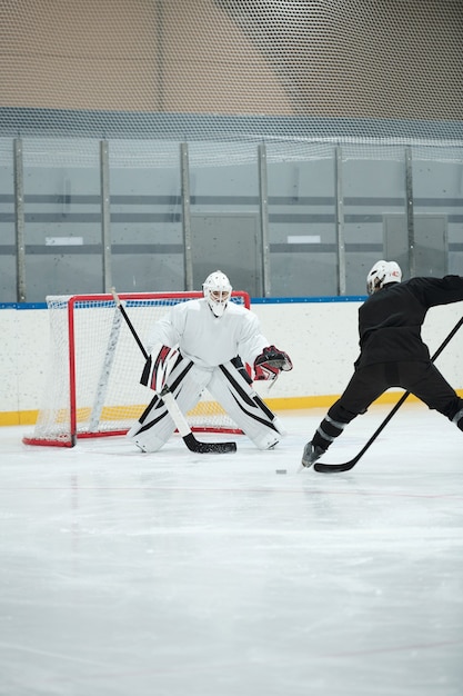 Jugador de hockey en uniforme deportivo blanco, casco protector, guantes y patines de pie en la pista frente a su rival y va a disparar el disco durante el juego