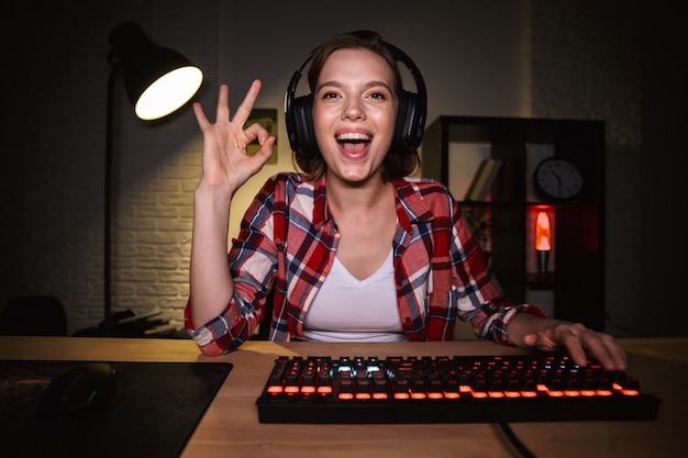 Jugador de mujer emocionada sentada en la mesa, jugando juegos en línea en una computadora en el interior, celebrando el éxito, mostrando ok