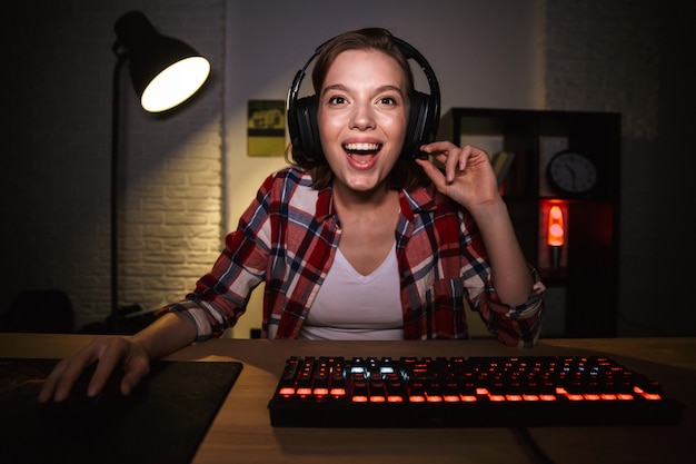 Jugador de mujer emocionada sentado en la mesa, jugando juegos en línea en una computadora en el interior, celebrando el éxito