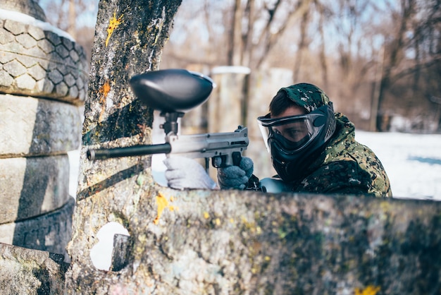 Jugador de paintball con máscara de protección disparando al enemigo, vista posterior, batalla de bosque de invierno, paintball. Juego de deporte extremo