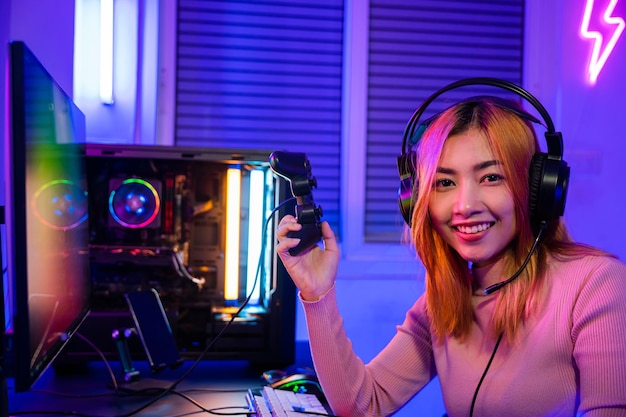 Un jugador sonriente que usa un controlador de joystick para un torneo juega un videojuego en línea con luz de neón de la computadora. Una mujer usa auriculares de juego jugando a juegos de esports en vivo. Consola mirando a la cámara.