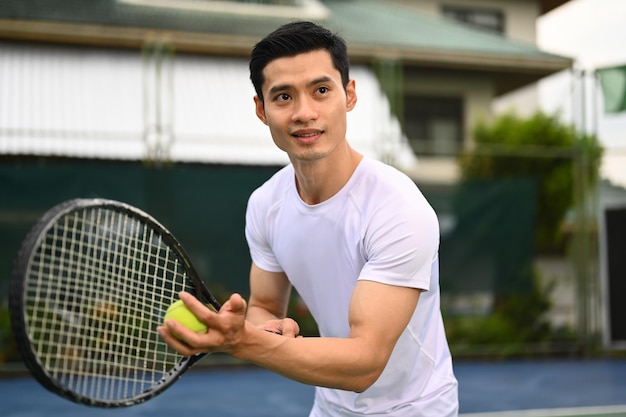 Jugador de tenis masculino asiático enfocado que sirve pelota de tenis durante un partido en cancha abierta Entrenamiento deportivo y concepto de vida activa