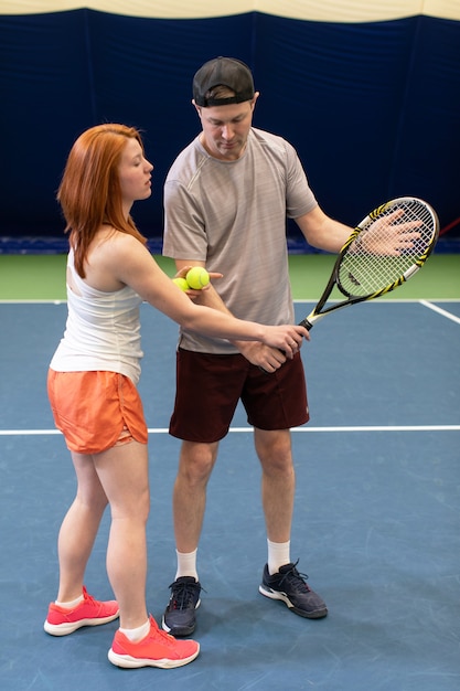 Jugador de tenis recibiendo instrucciones del entrenador sobre cómo jugar en una cancha cubierta. Instructora explica la técnica de golpear a su alumno