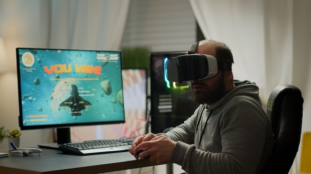 Jugador de videojuegos con casco de realidad virtual levantando la mano después de ganar la competencia de disparos espaciales. Jugador profesional profesional que juega a videojuegos en línea con nuevos gráficos en una computadora potente desde la sala de juegos