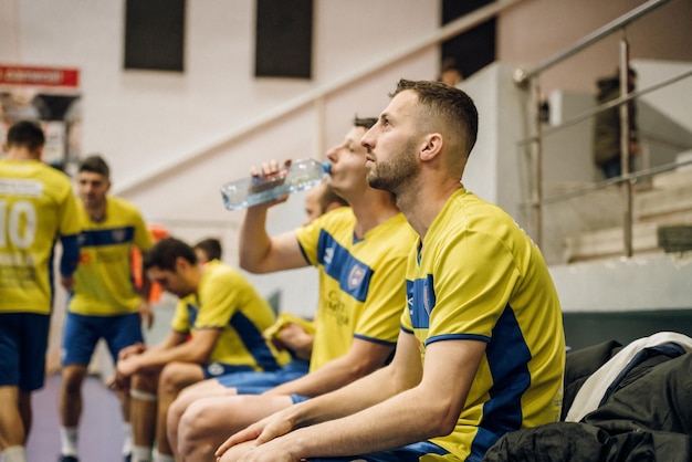 Jugadores sentados en un gimnasio, bebiendo agua y vistiendo camisetas azules y amarillas