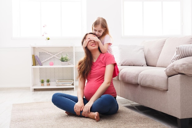 Jugando al escondite junto con la mamá embarazada, sentada en el suelo en casa. Concepto de amor, maternidad y cuidado familiar, espacio de copia.