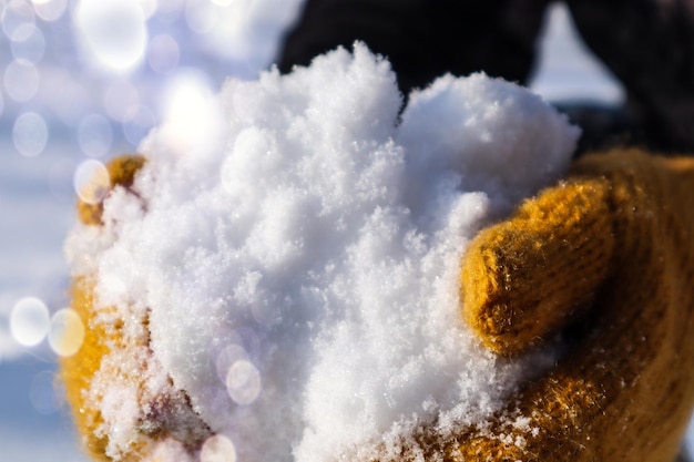 Jugando en la nieve, las manos con mitones amarillos sostienen la nieve para hacer un bokeh de primer plano de bola de nieve