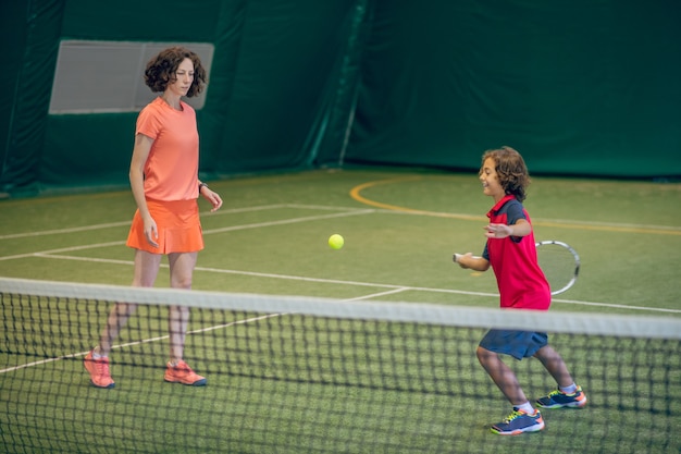 Jugando tenis. Entrenador femenino en ropa brillante enseñando a un niño a jugar al tenis en un gimnasio