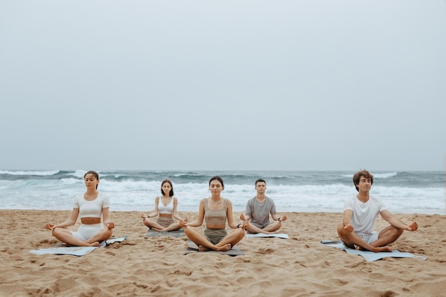 Jugend-Strand-Yoga-Gruppe von Frauen und Männern, die am Strand am Meeresufer meditieren und auf Matten sitzen