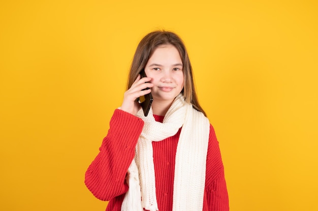 Jugendlich Mädchen im warmen weißen gestrickten Schal, der auf dem Handy lokalisiert auf gelbem Hintergrund spricht