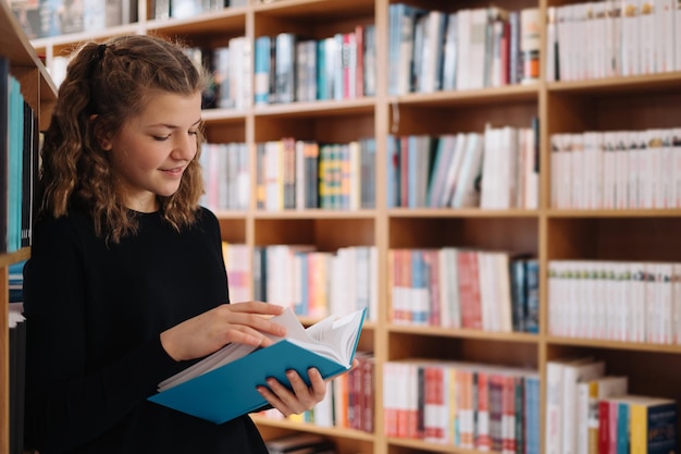 Jugendlich Mädchen unter einem Stapel Bücher. Ein junges Mädchen liest ein Buch mit Regalen im Hintergrund. Sie ist von Bücherstapeln umgeben. Tag buchen.