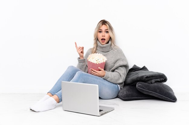 Jugendlichblondine, die Popcorn beim Aufpassen eines Films auf dem Laptop überrascht und Seite zeigend essen