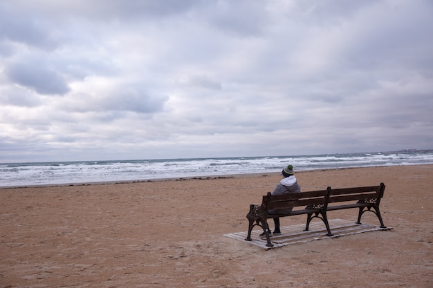 Jugendlicher, der auf einer Bank auf einem leeren Strand, bewölkter Tag sitzt. Das Konzept der Einsamkeit.