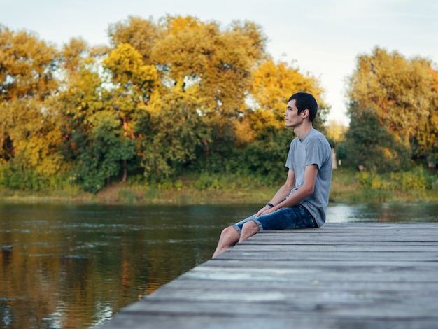 Jugendlicher sitzt auf einer Holzbrücke am Fluss im Freien und schaut im Gefolge des abreisenden Sommers. Letzte Sommertage