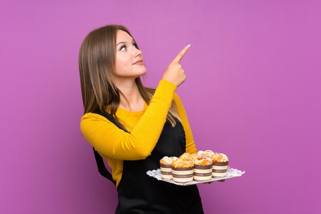 Jugendlichmädchen, das viele verschiedene Minikuchen über der lokalisierten purpurroten Wand zeigt mit dem Zeigefinger eine großartige Idee hält