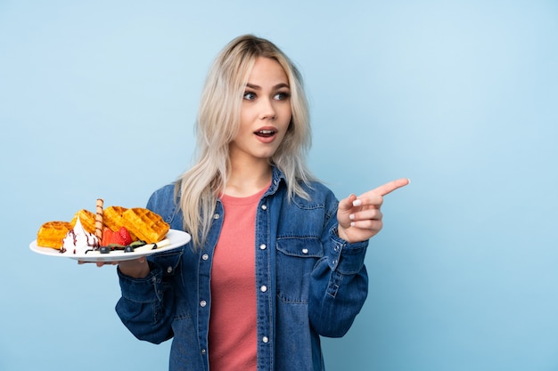 Jugendlichmädchen, das Waffeln über der blauen Wand überrascht hält und Finger auf die Seite zeigt