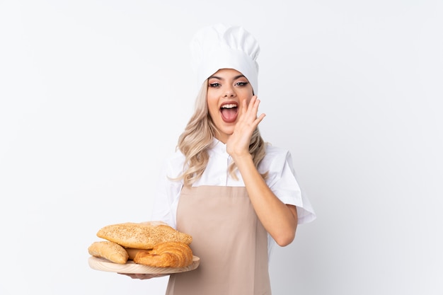 Jugendlichmädchen in der Chefuniform. Der weibliche Bäcker, der eine Tabelle mit einigen Broten über lokalisiertem weißem Hintergrund schreit mit dem breiten Mund hält, öffnen sich