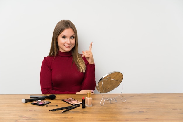 Jugendlichmädchen mit Make-uppalette und Kosmetik in einer Tabelle zeigend mit dem Zeigefinger eine großartige Idee