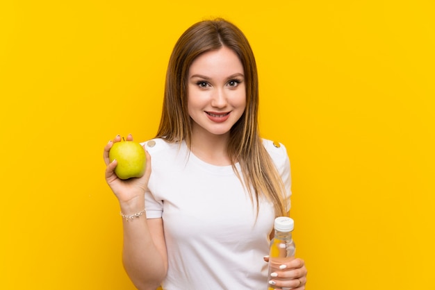 Jugendlichmädchen über gelber Wand mit einem Apfel und einer Flasche Wasser