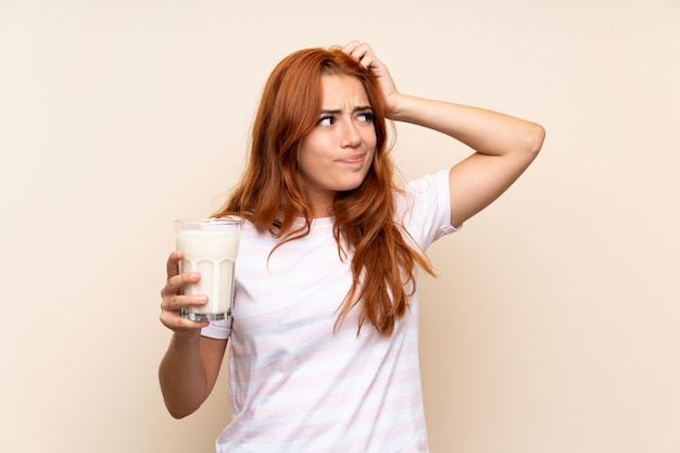 Jugendlichrothaarigemädchen, das ein Glas Milch über lokalisiertem Haben von Zweifeln und mit verwirren Gesichtsausdruck hält