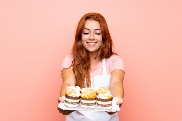 Jugendlichrothaarigemädchen, das viele verschiedene Minikuchen über lokalisiertem rosa Hintergrund hält