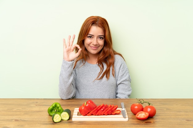 Jugendlichrothaarigemädchen mit Gemüse in einer Tabelle, die ein okayzeichen mit den Fingern zeigt