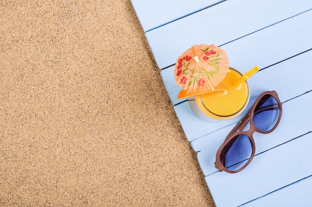 Jugo de naranja cítrico en un vaso y gafas de sol en tableros azules en la playa