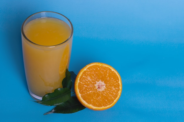 Jugo de naranja fresco en un vaso con fruta cortada por la mitad y en rodajas por hojas verdes aisladas sobre un fondo azul, vista superior, copie el espacio.