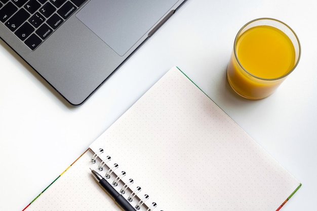 Jugo de naranja en el lugar de trabajo. Computadora portátil, vidrio de zumo de naranja y cuaderno en el escritorio blanco.