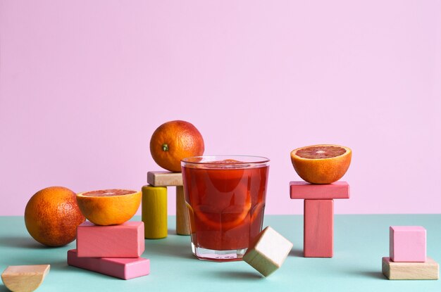 Jugo de naranja en un vaso con formas geométricas podims y naranjas sobre ellos sobre fondo rosa y menta