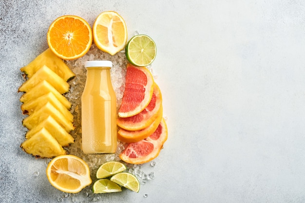 Jugo de piña fresca en frasco de vidrio con hielo y variedad en rodajas maduras de color frutas tropicales pomelo, naranja, lima, limón, composición plana, sobre fondo de piedra gris claro.